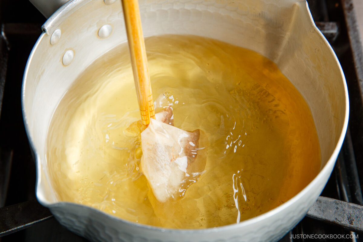 A wooden spatula stirs a clear golden liquid, likely dashi broth, in a silver pot on a stovetop.