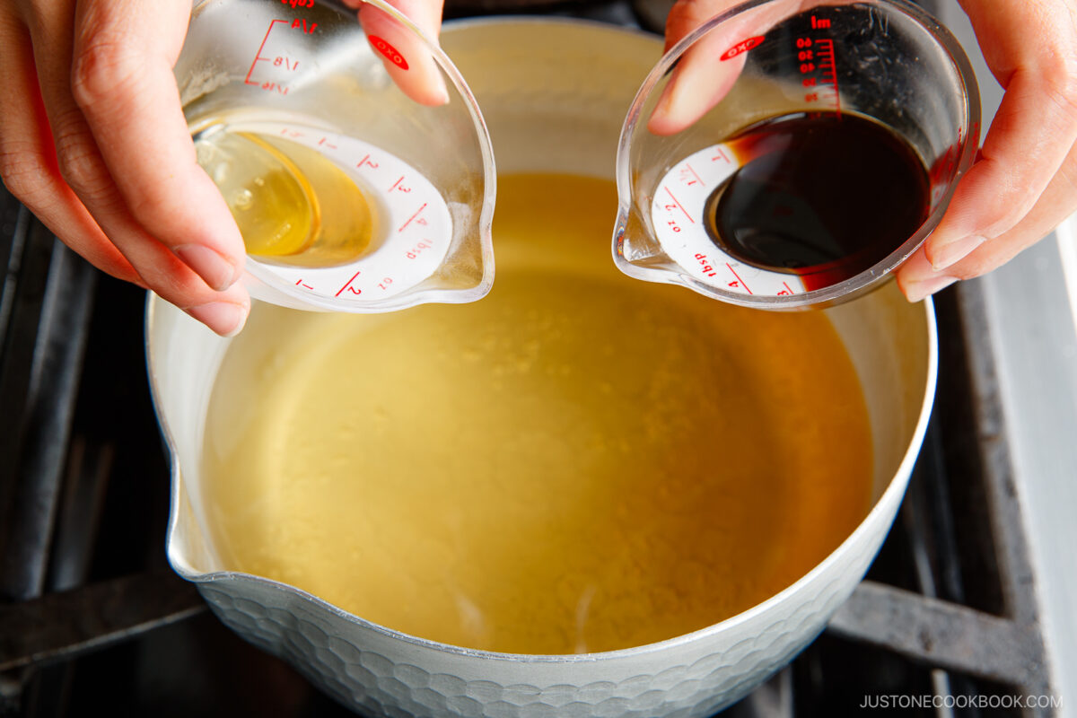 Two hands holding measuring cups, one with a light liquid and one with a dark liquid, over a pot of light-colored broth on a stove. The liquids are about to be poured into the pot.