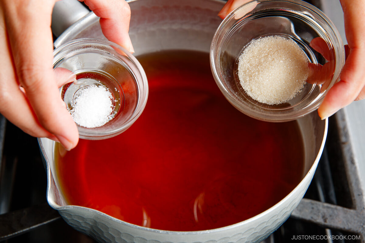 Two hands hold small glass bowls of salt and sugar over a pot filled with red liquid on a stovetop, preparing to add the ingredients.
