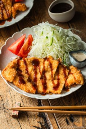 A plate of crispy breaded chicken cutlets drizzled with sauce, served with shredded cabbage, tomato wedges, and dipping sauce. Chopsticks rest in front of the plate on a wooden table.