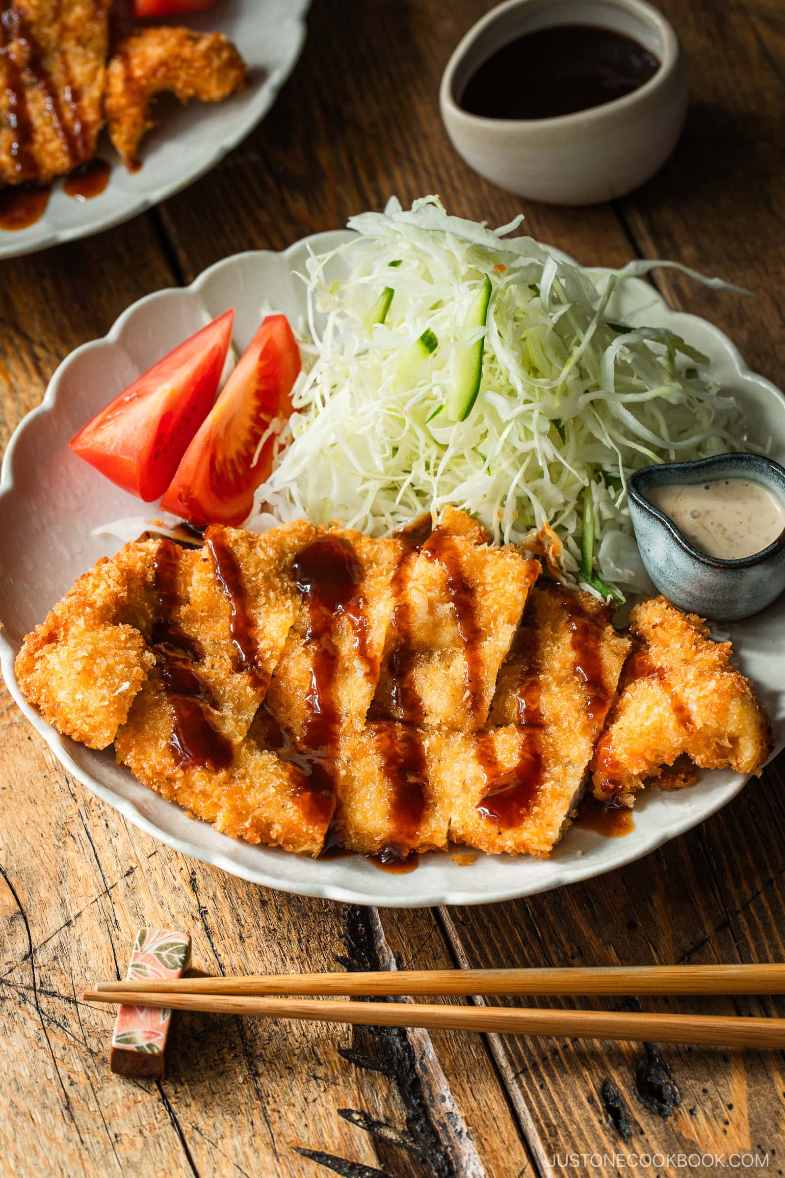 A plate of crispy breaded chicken cutlets drizzled with sauce, served with shredded cabbage, tomato wedges, and dipping sauce. Chopsticks rest in front of the plate on a wooden table.