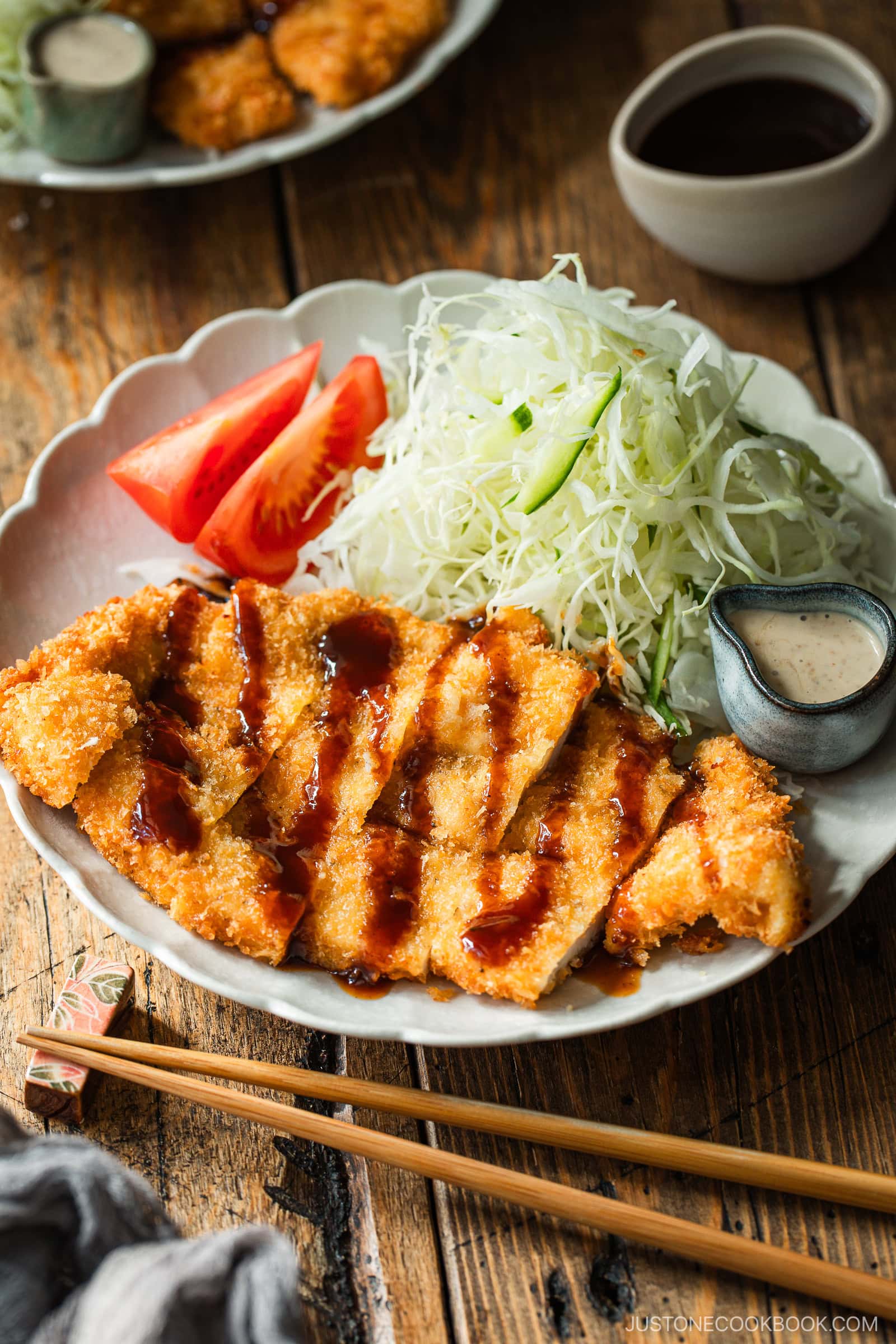 A plate of Japanese tonkatsu (breaded pork cutlet) sliced and drizzled with sauce, served with shredded cabbage, tomato wedges, dipping sauces, and wooden chopsticks on a rustic table.