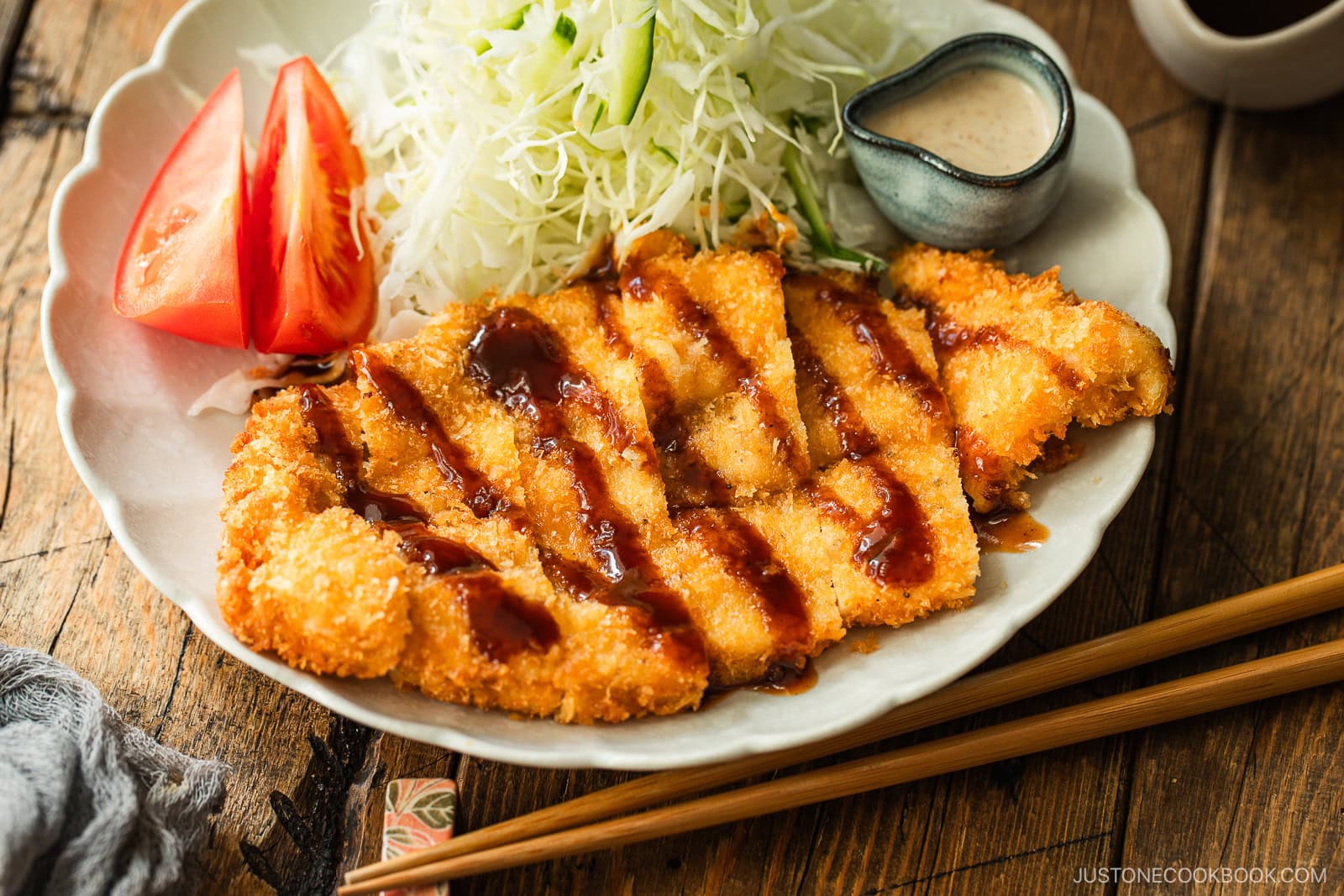 A plate of crispy breaded chicken cutlets drizzled with sauce, served with shredded cabbage, tomato wedges, chopsticks, and a small dish of creamy dressing on a wooden table.