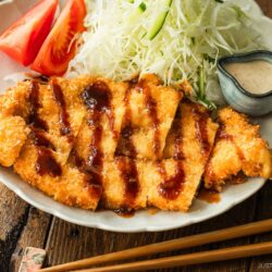 A plate of crispy breaded chicken cutlets drizzled with sauce, served with shredded cabbage, tomato wedges, chopsticks, and a small dish of creamy dressing on a wooden table.