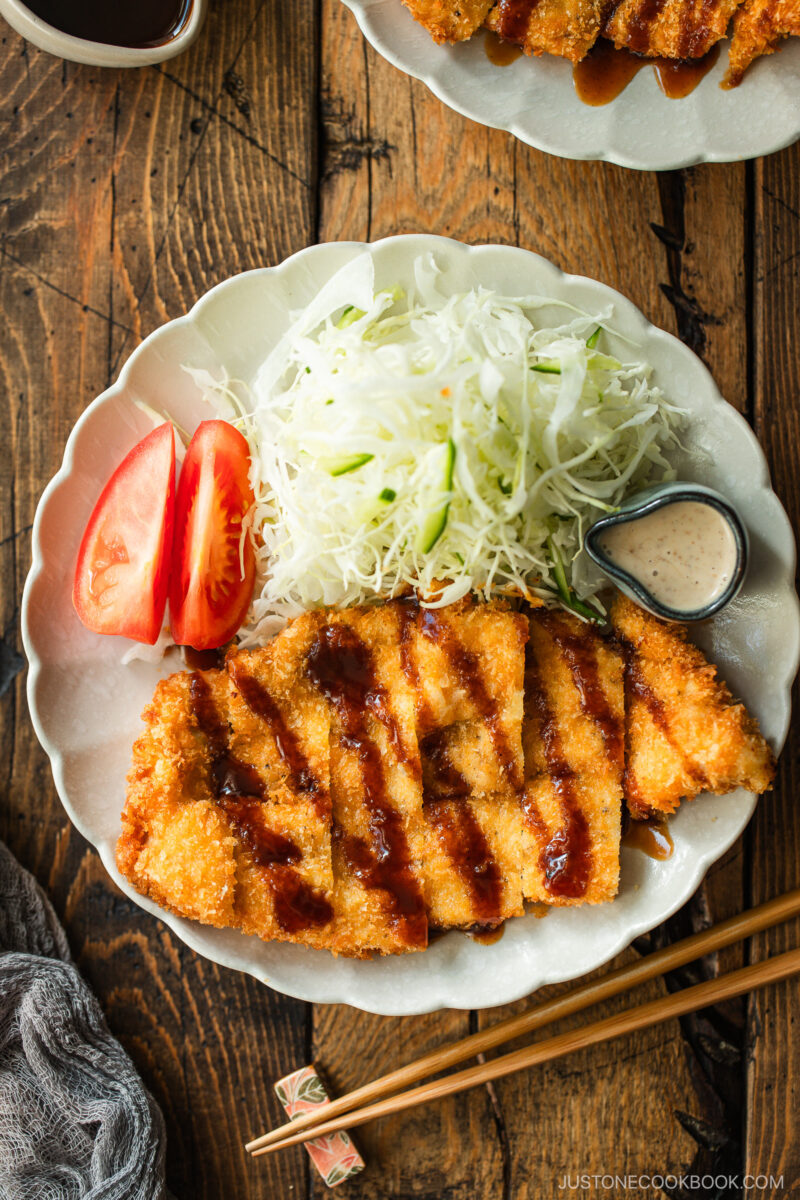 A plate with sliced breaded chicken cutlet drizzled with sauce, shredded cabbage, tomato wedges, and a small container of creamy dressing, served with chopsticks on a wooden table.
