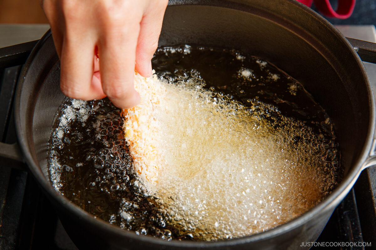 A hand lowers a breaded food item into a pot of hot oil, causing bubbles to form as the item begins to fry.