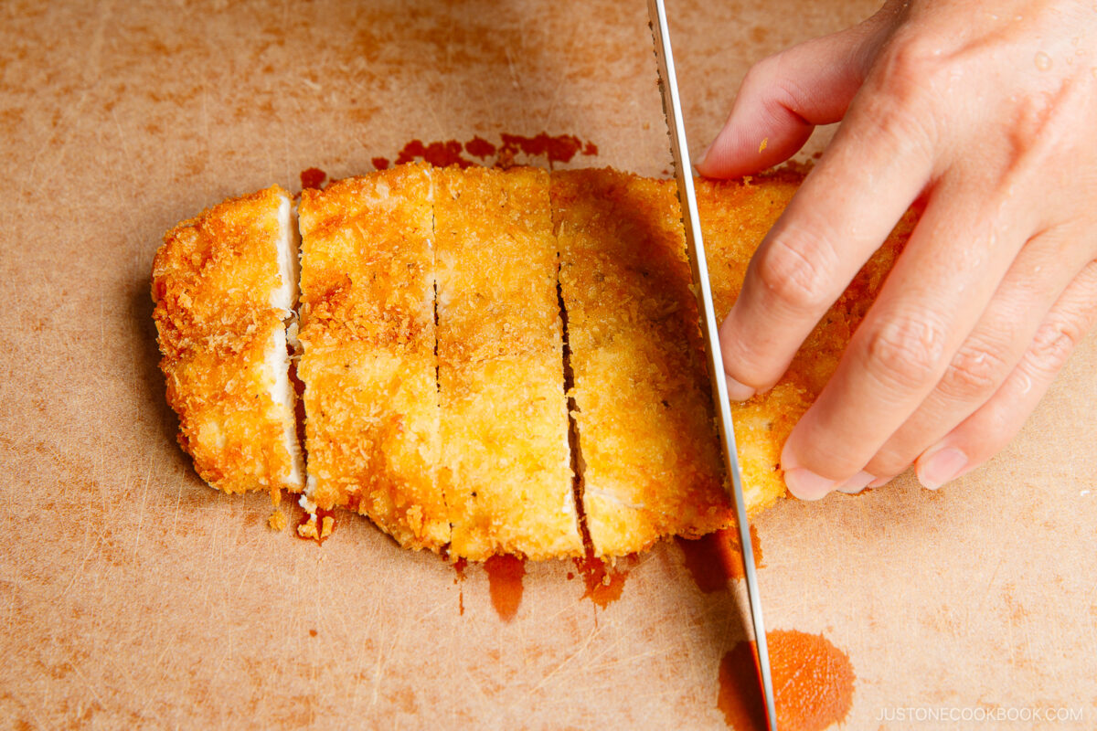 A hand slices a piece of golden, crispy breaded chicken cutlet on a wooden cutting board, with a sharp knife creating even pieces.