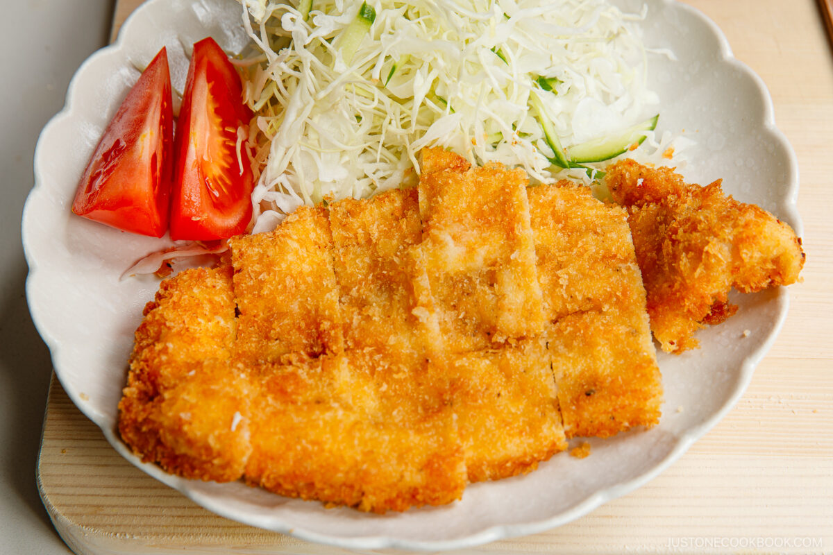 A plate with crispy breaded chicken cutlet, sliced and served with shredded cabbage, cucumber, and tomato wedges on the side.