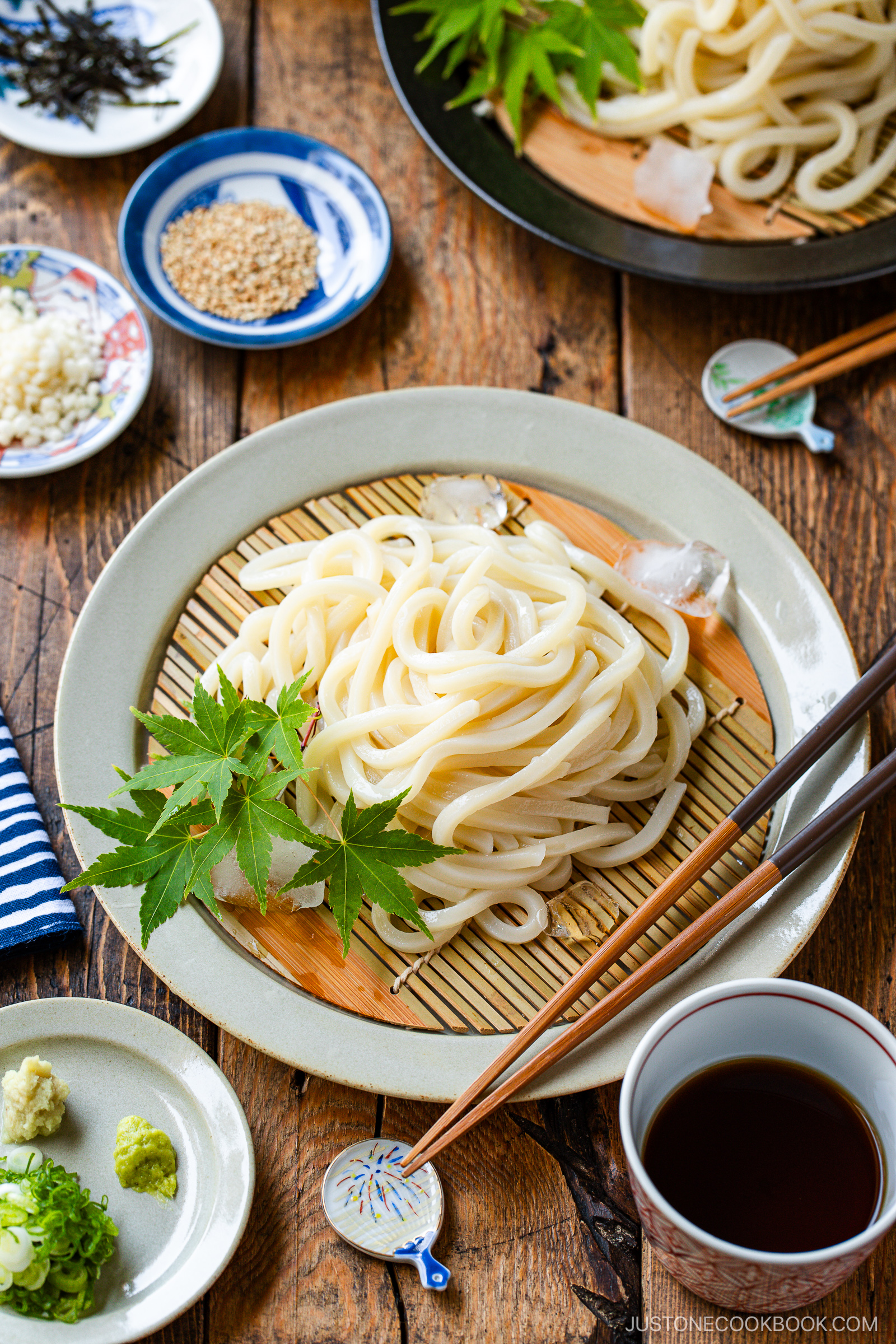 A plate of chilled udon noodles on a bamboo mat, garnished with green maple leaves, served with dipping sauce, wasabi, green onions, ginger, and sesame seeds on a rustic wooden table.