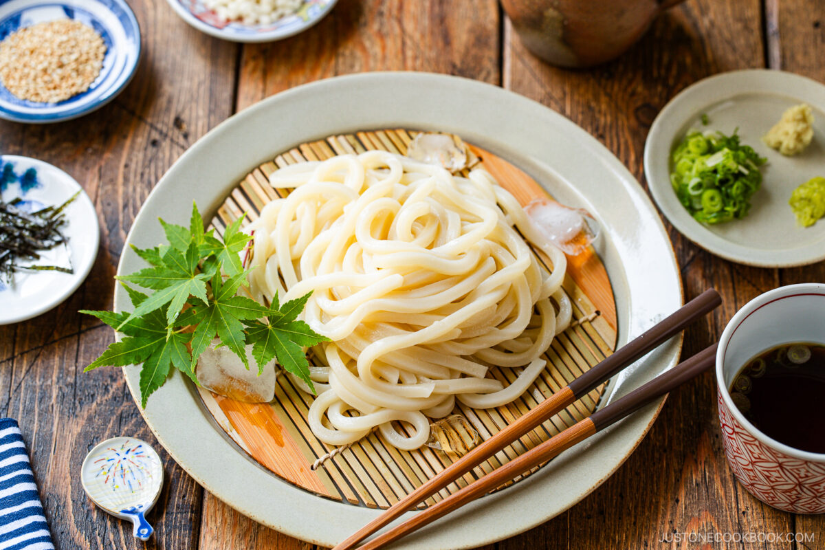 A plate of chilled udon noodles served on a bamboo mat with ice cubes, garnished with maple leaves. Sides of sliced green onions, wasabi, shredded nori, and dipping sauce are arranged around the plate on a wooden table.