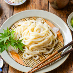 A plate of chilled udon noodles served on a bamboo mat with ice cubes, garnished with maple leaves. Sides of sliced green onions, wasabi, shredded nori, and dipping sauce are arranged around the plate on a wooden table.