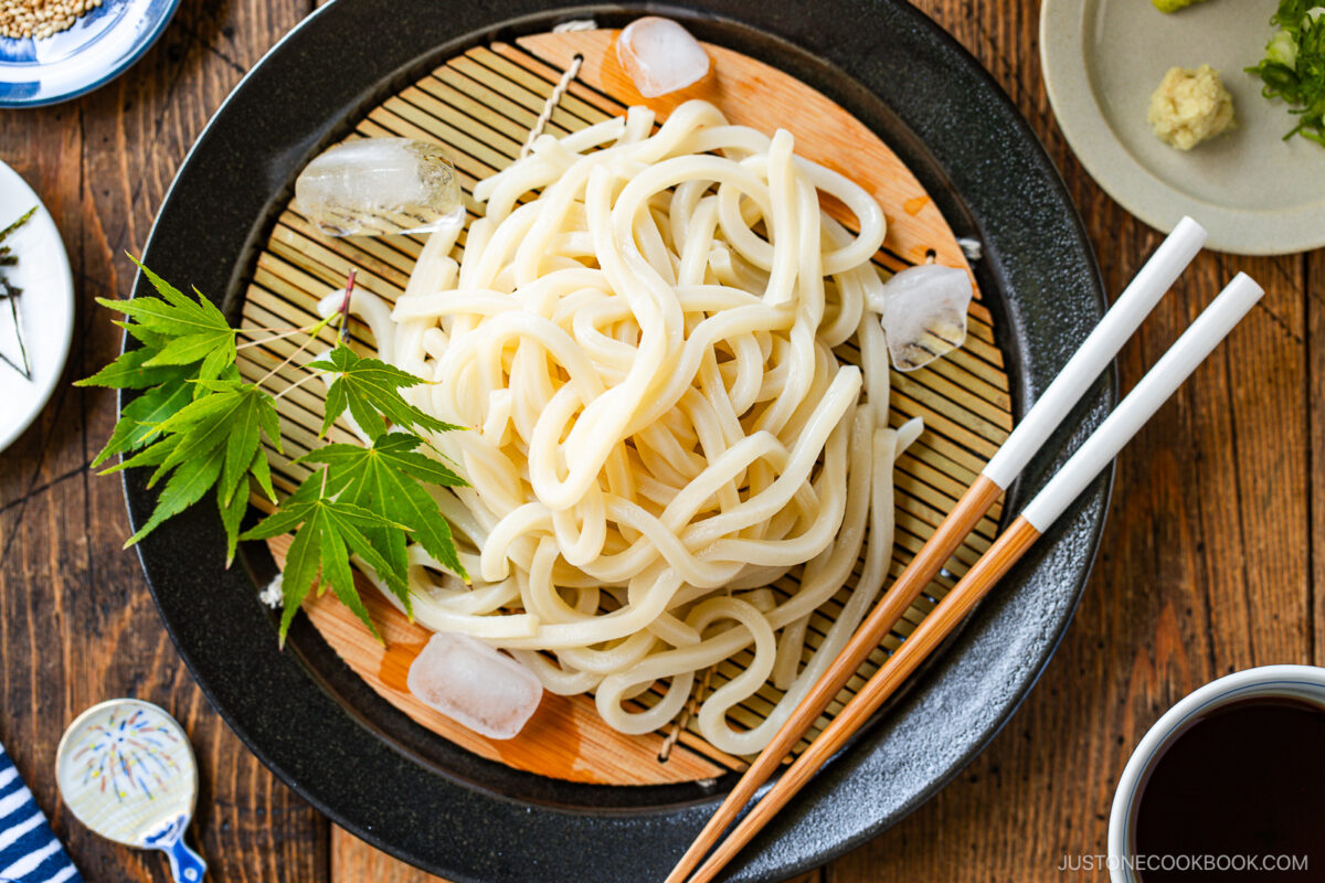 A plate of cold udon noodles served on a bamboo mat with ice cubes, garnished with green maple leaves. Chopsticks are placed on the side, and small dishes surround the main plate on a wooden table.