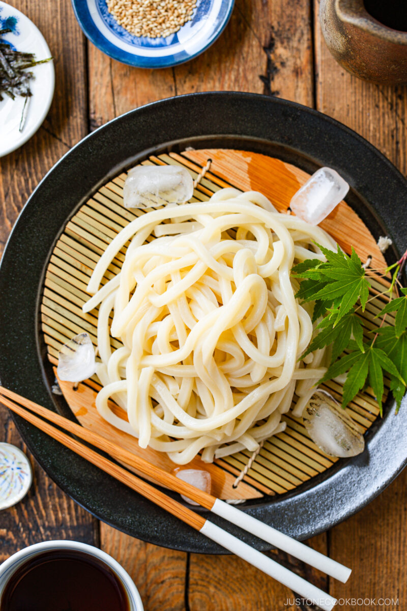 A plate of chilled udon noodles on a bamboo mat with ice cubes, garnished with a green maple leaf. Wooden chopsticks rest on the plate, and bowls of dipping sauce and sesame seeds are nearby on a wooden table.