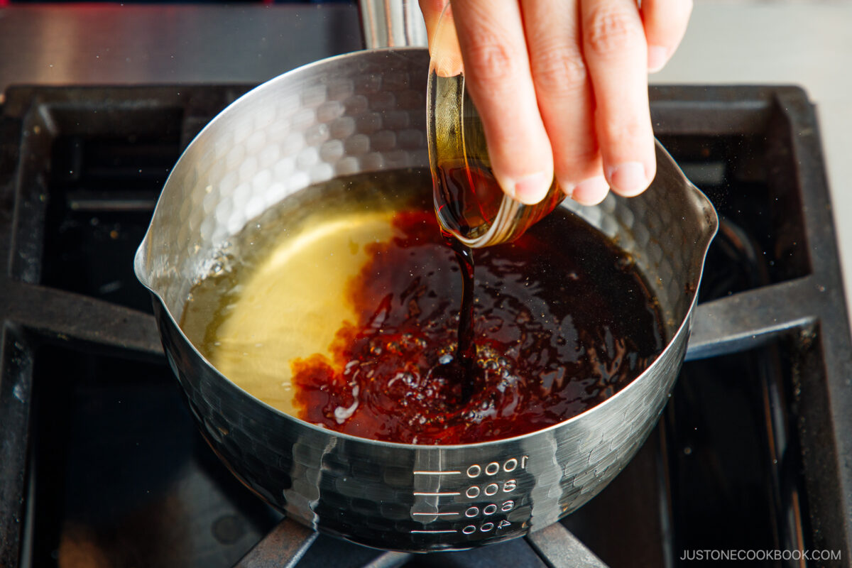 A hand pours a dark liquid from a small glass bowl into a saucepan with a light-colored liquid, on a stovetop.