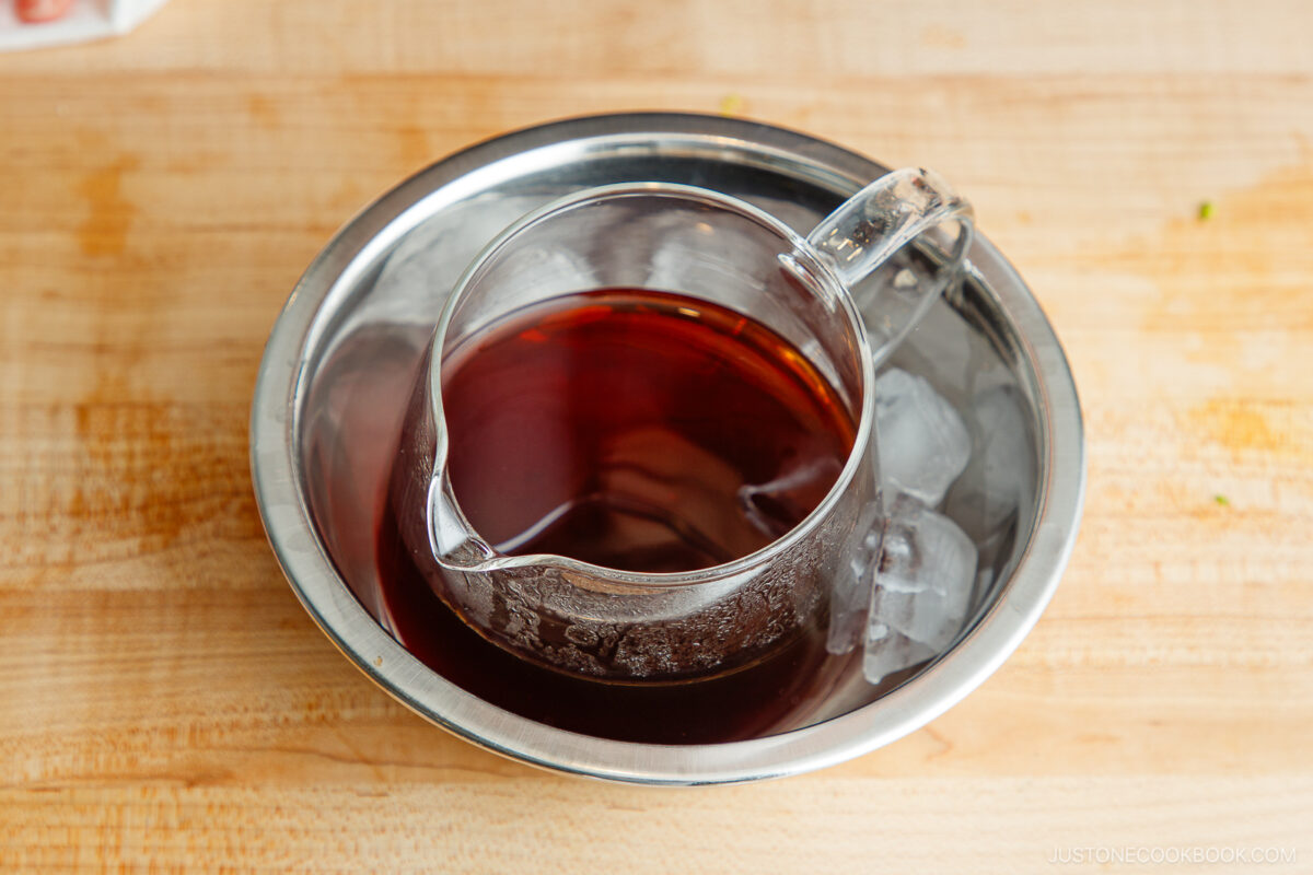 A glass measuring cup filled with dark liquid is placed in a metal bowl with ice cubes on a wooden surface, cooling the liquid.