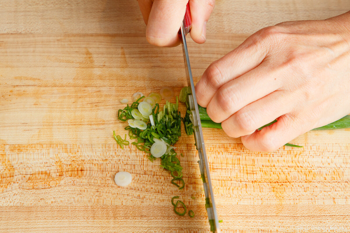 A close-up of hands slicing green onions on a wooden cutting board with a knife. Thin, round slices of the green onion are scattered on the board.