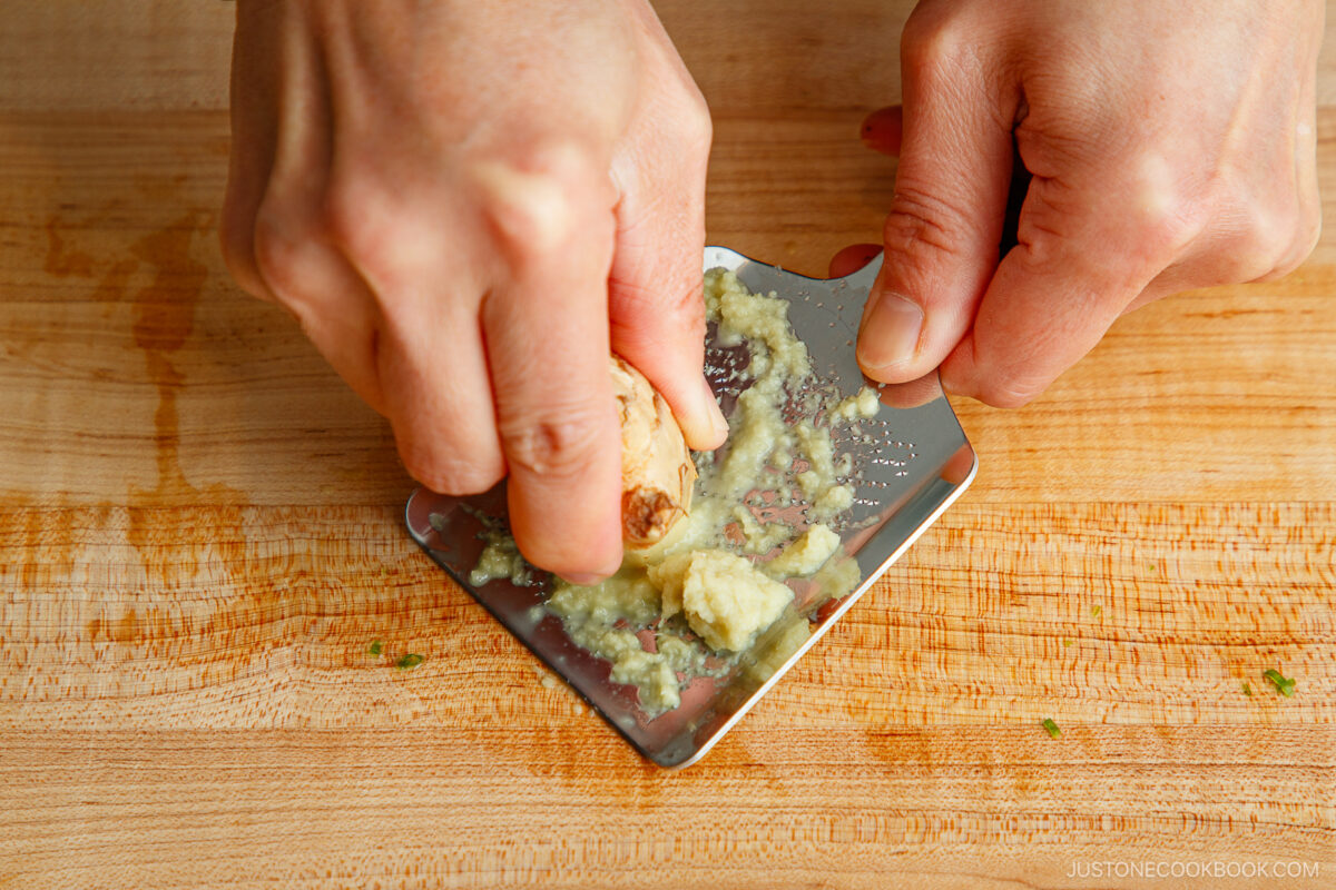A close-up of udon noodles being boiled in a large stainless steel pot filled with water on a stovetop. The noodles are partially submerged and beginning to separate as they cook.