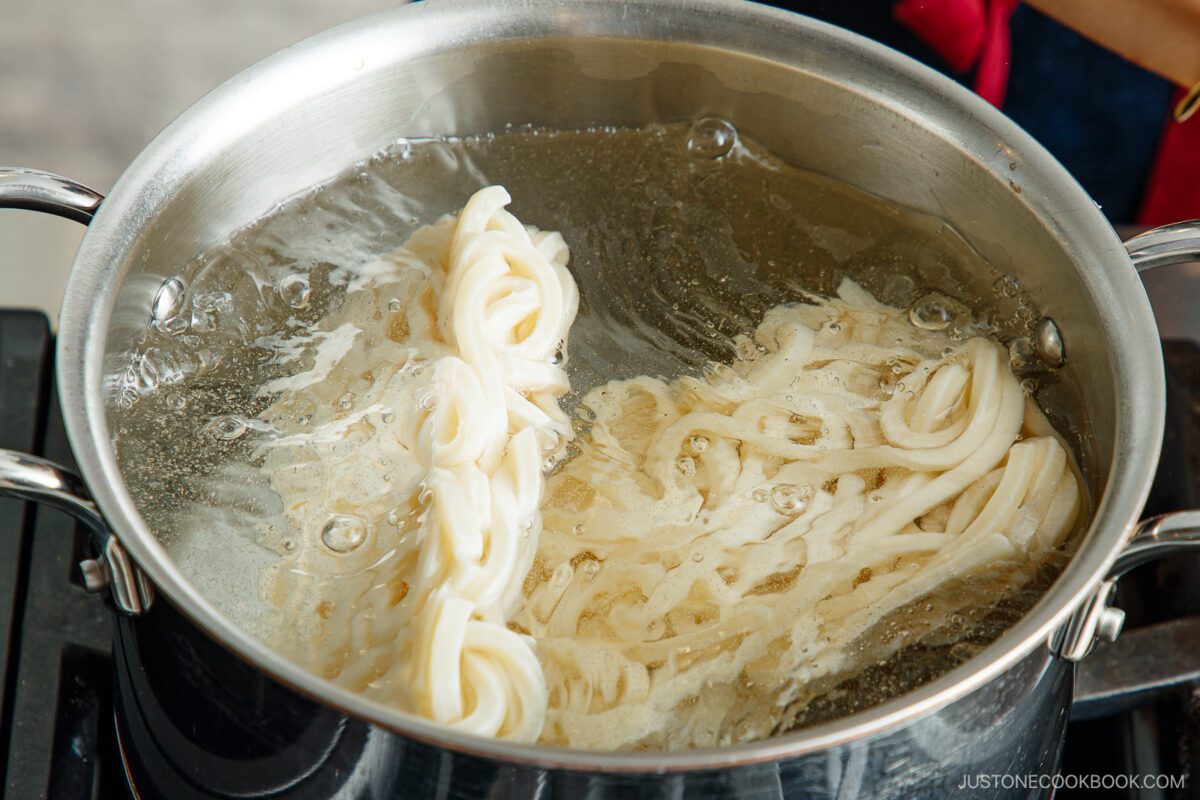 A close-up of udon noodles being boiled in a large stainless steel pot filled with water on a stovetop. The noodles are partially submerged and beginning to separate as they cook.