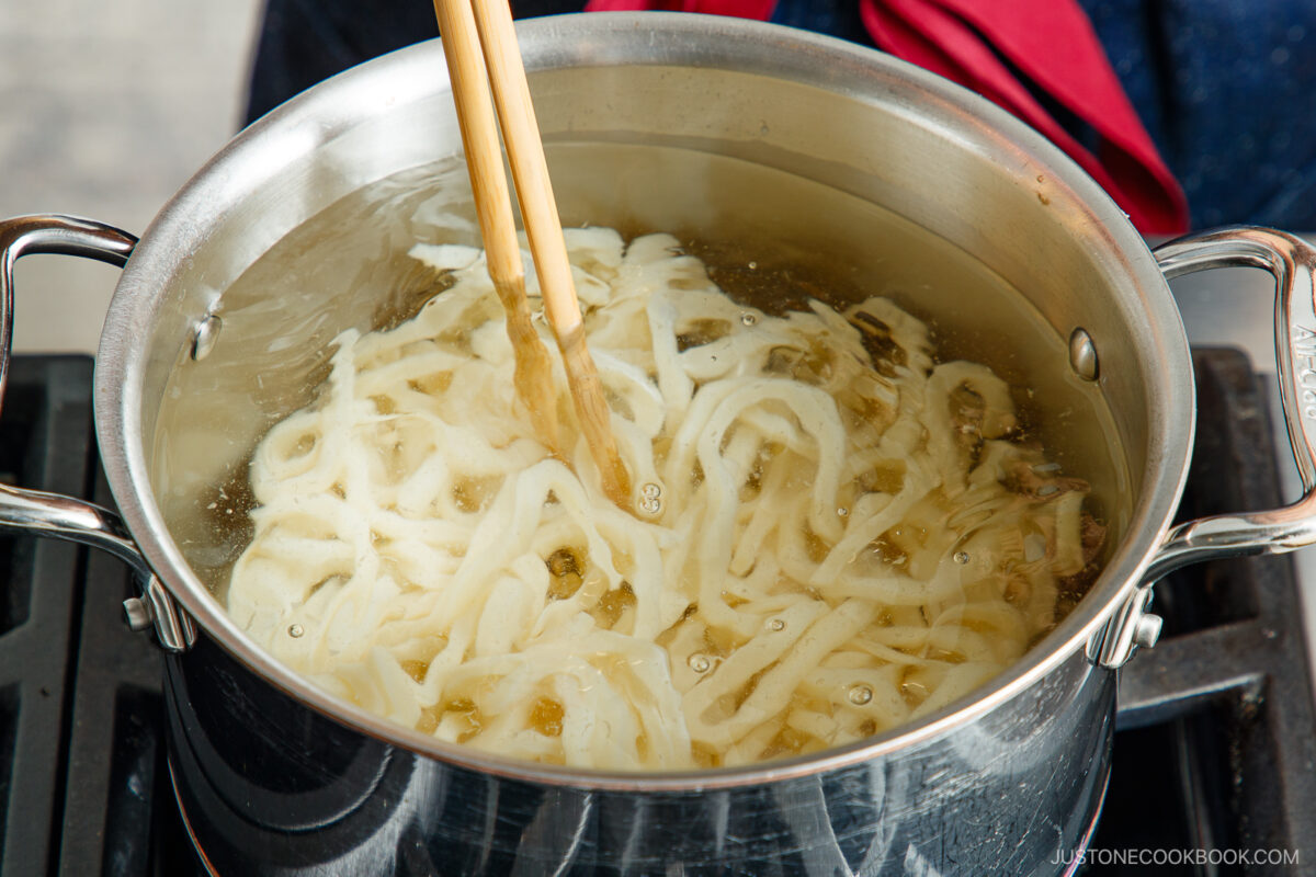 A pot of simmering clear broth is being stirred with a wooden spoon on a stovetop. The liquid is light golden in color, with bubbles forming as it heats.