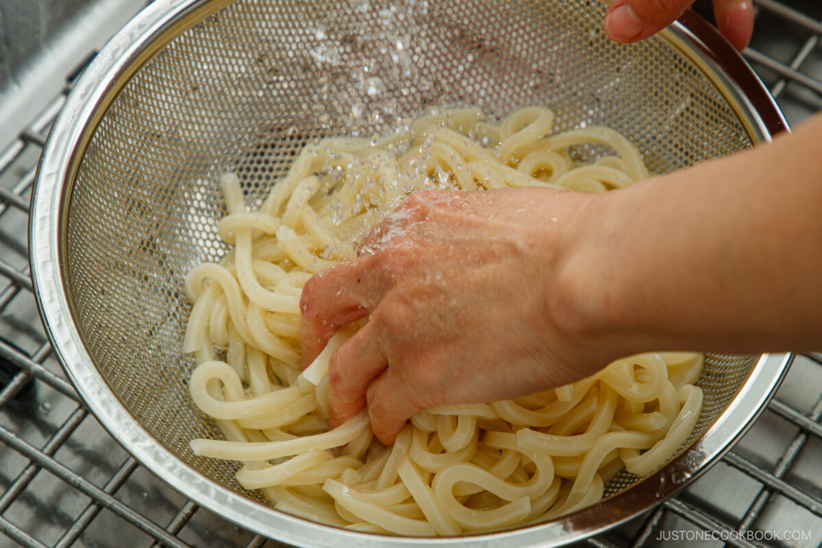 A hand rinsing cooked udon noodles under running water in a metal colander placed in a sink.
