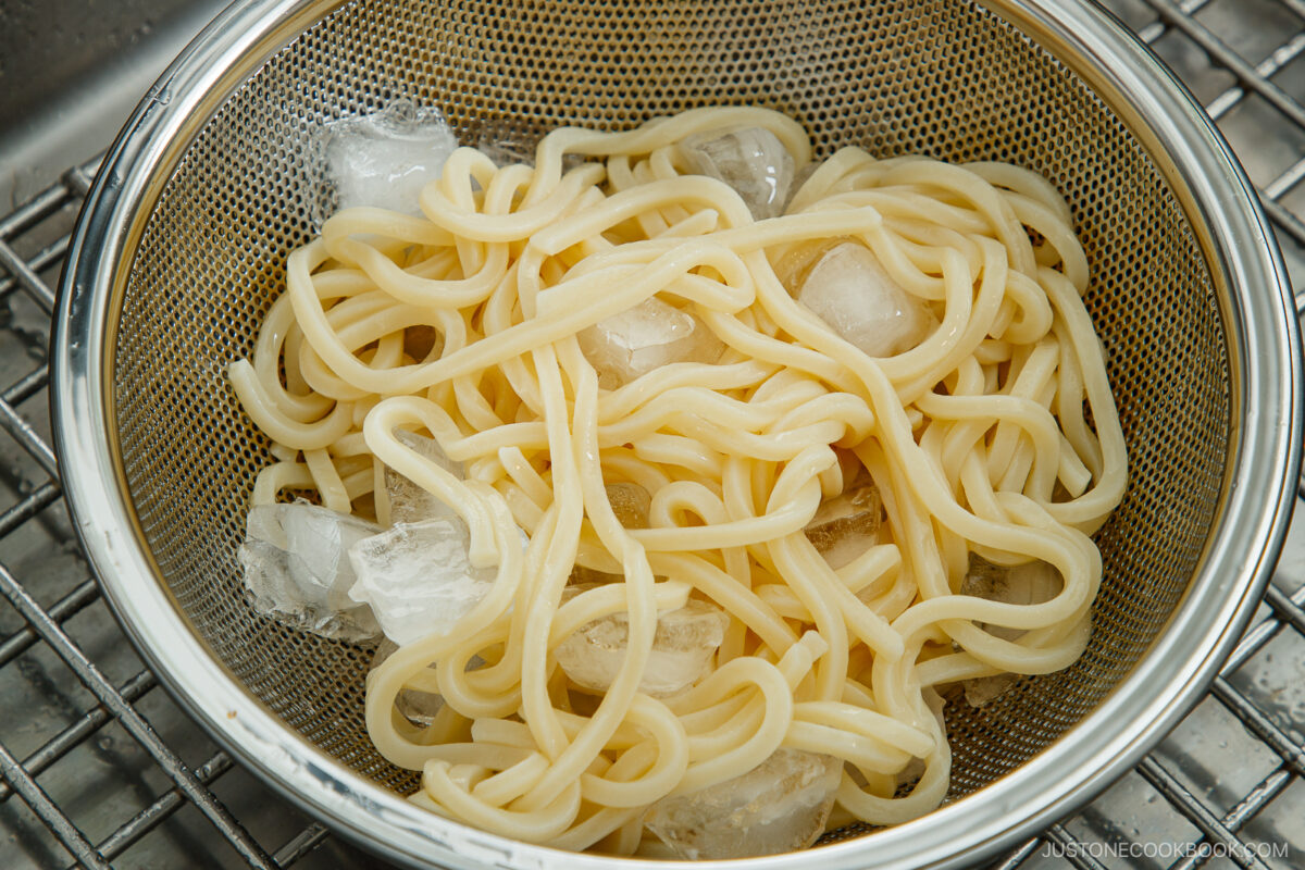 A metal colander contains cooked udon noodles with ice cubes, sitting on a sink rack, likely being cooled or rinsed.