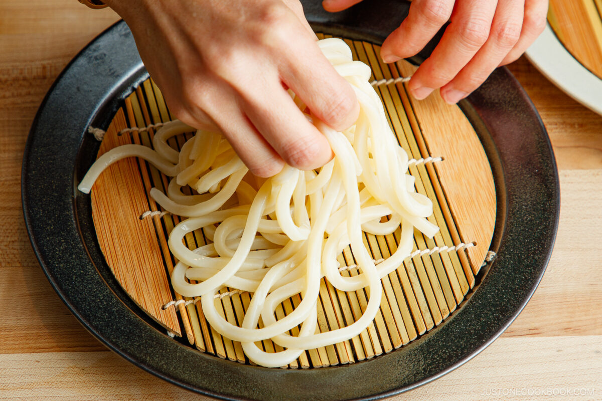 A person arranges cooked udon noodles on a bamboo mat placed on a round black plate.