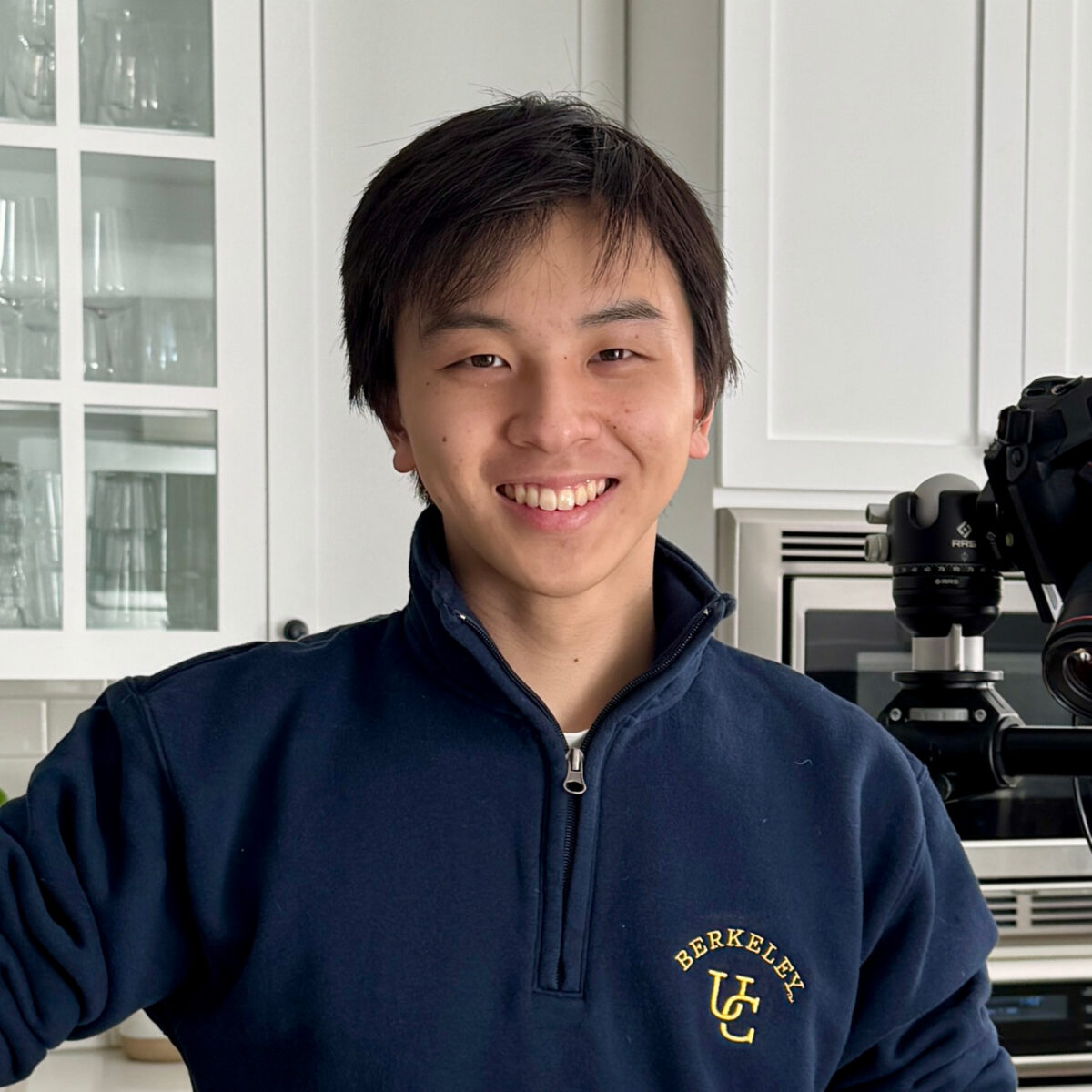 Young Asian man smiling in a kitchen with a professional camera and microscope, wearing a navy Berkeley sweatshirt.