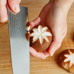 A person uses a knife to carve a decorative star pattern into the cap of a shiitake mushroom on a wooden cutting board.