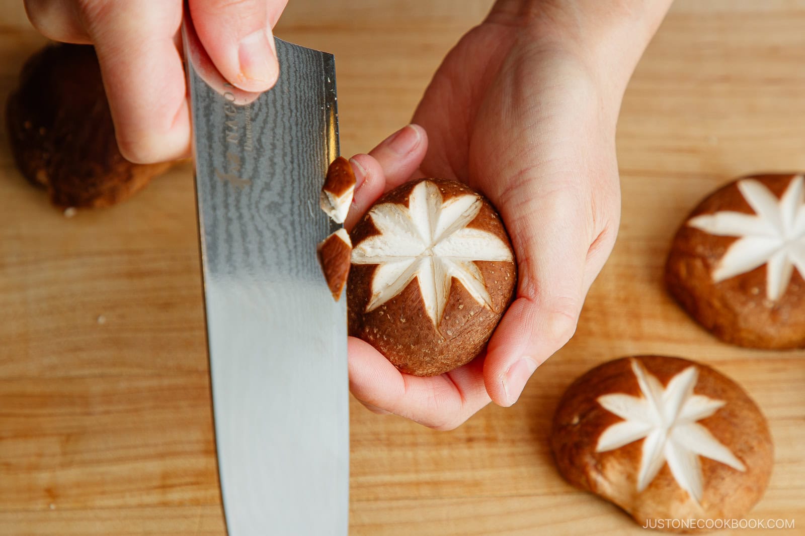 A person uses a knife to carve a decorative star pattern into the cap of a shiitake mushroom on a wooden cutting board.