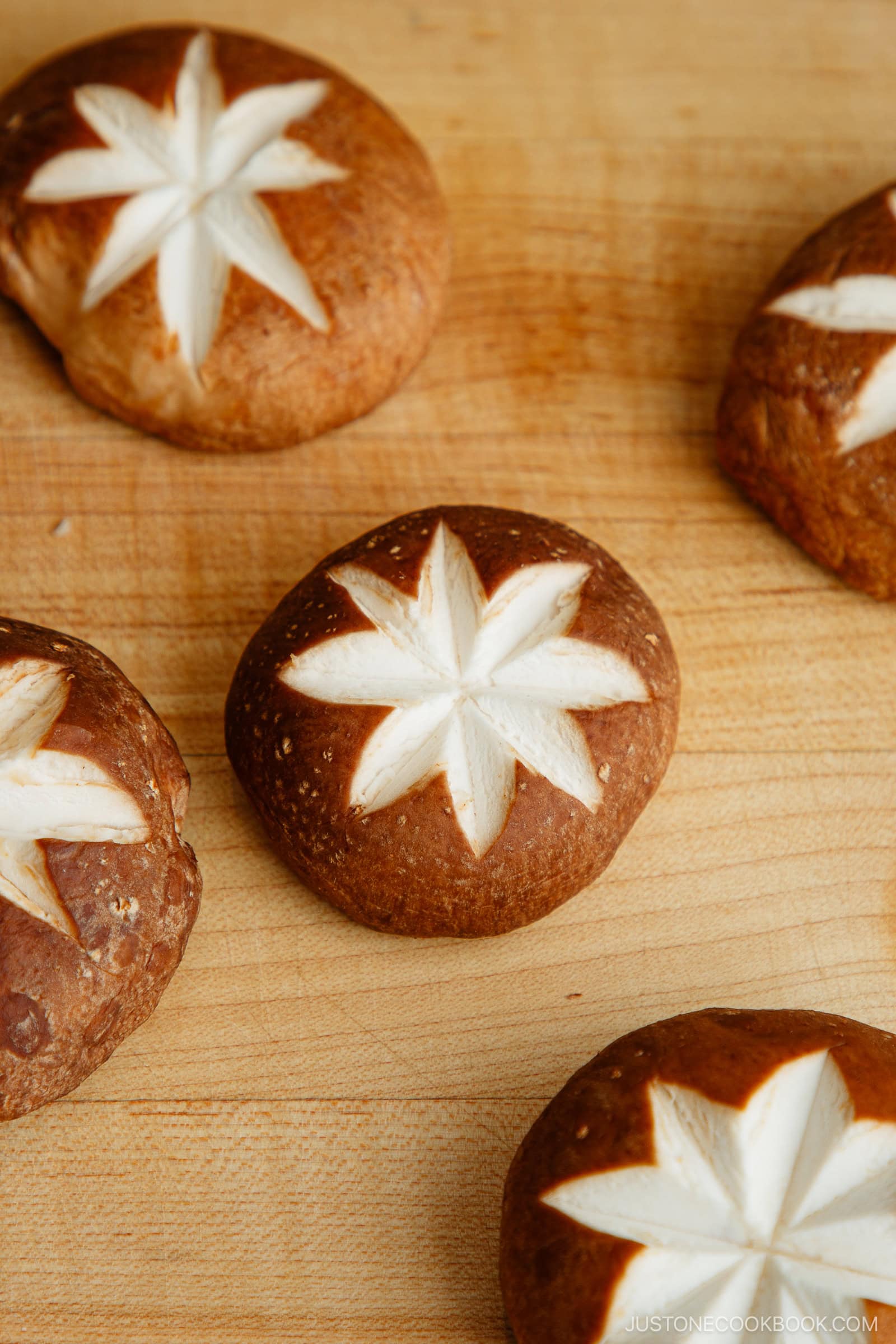 Five shiitake mushrooms with decorative star-shaped cuts on their caps are arranged on a light wooden surface.
