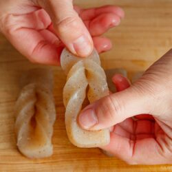 Two hands twist a piece of grey konnyaku (yam cake) into a decorative knot on a wooden surface, with additional twisted konnyaku pieces in the background.
