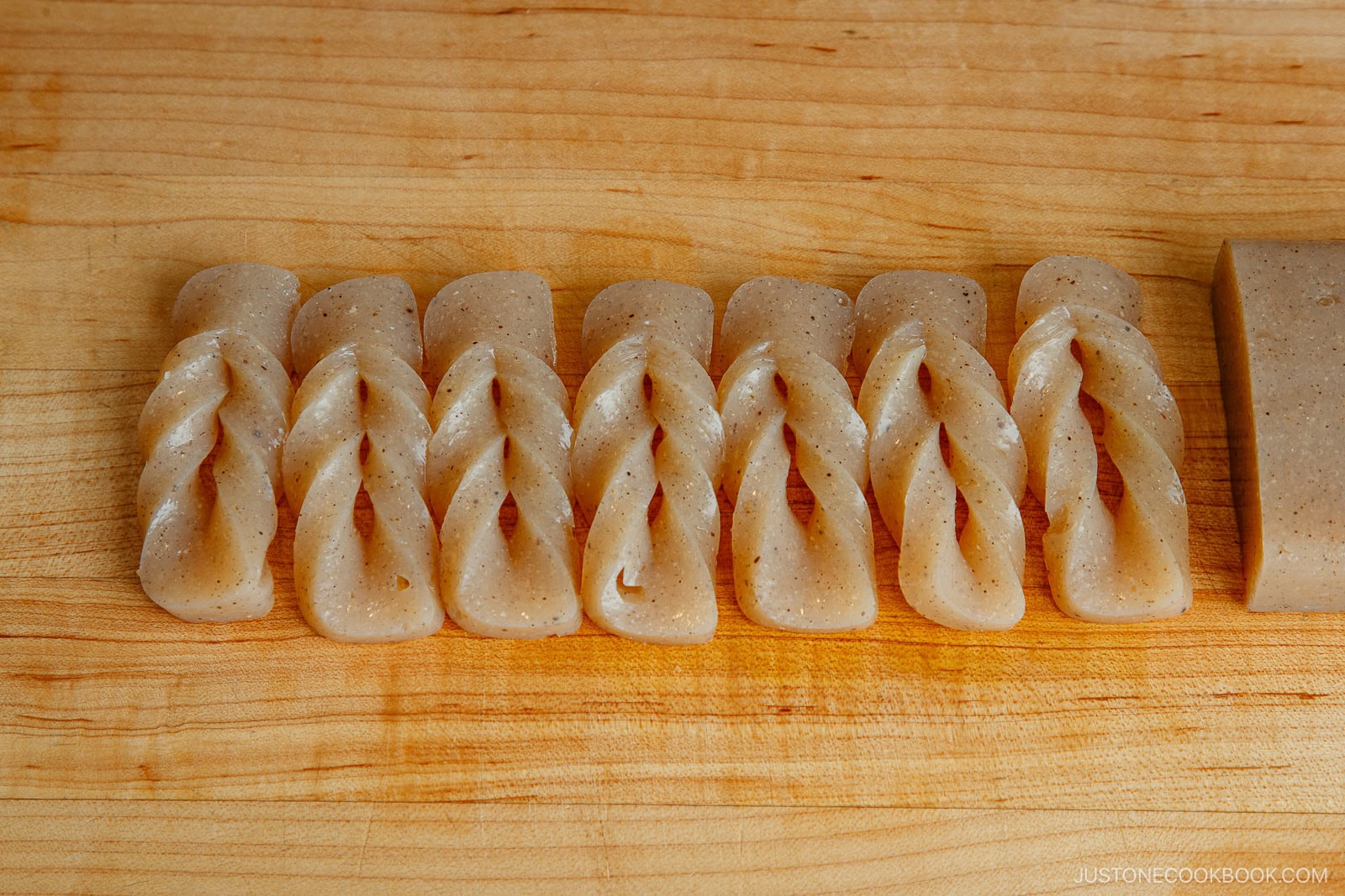 Eight pieces of twisted, translucent konnyaku (yam cake) are neatly lined up on a wooden cutting board, with an uncut block on the right side.