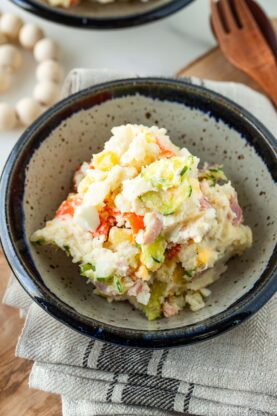 A bowl of Japanese potato salad with visible chunks of potato, carrot, cucumber, and ham, served in a rustic ceramic bowl on a folded cloth napkin beside a wooden fork and spoon.