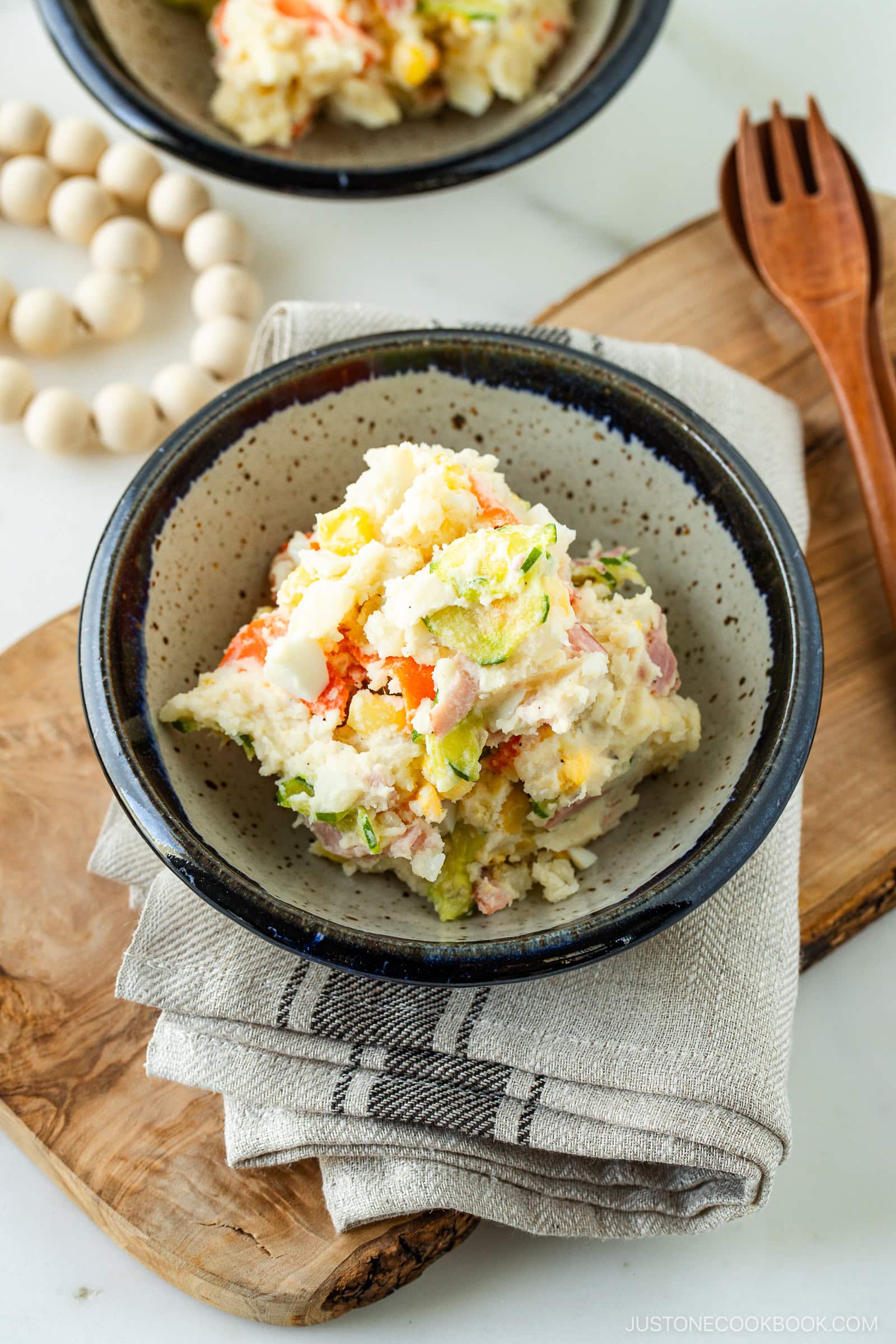 A bowl of Japanese potato salad with visible chunks of potato, carrot, cucumber, and ham, served in a rustic ceramic bowl on a folded cloth napkin beside a wooden fork and spoon.