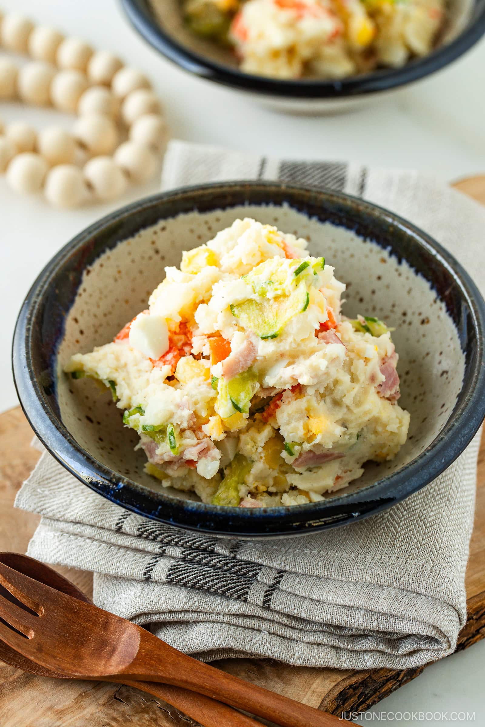 A bowl of Japanese potato salad with visible chunks of potato, carrot, cucumber, and ham, served in a rustic ceramic bowl on a folded cloth napkin beside a wooden fork and spoon.