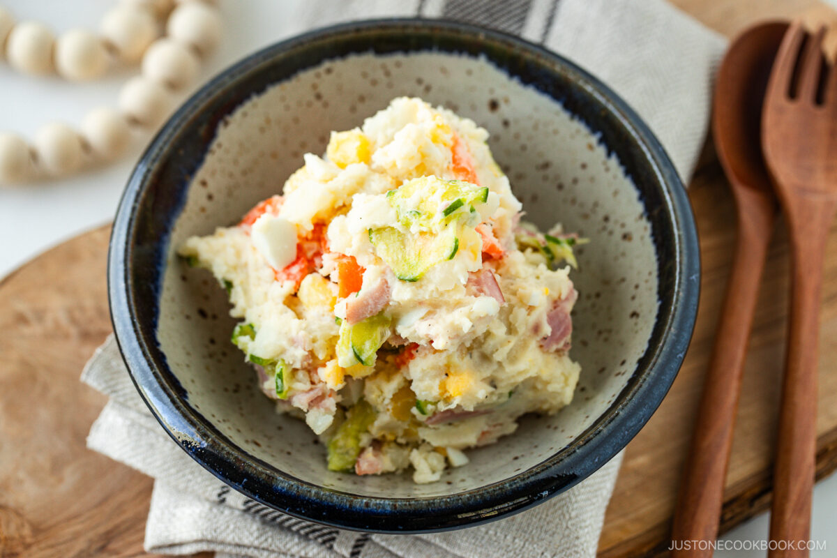 A bowl of Japanese potato salad with visible chunks of potato, ham, boiled egg, cucumber, and carrot, placed on a rustic plate beside wooden utensils and a beaded decoration.