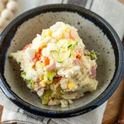 A bowl of Japanese potato salad with visible chunks of potato, ham, boiled egg, cucumber, and carrot, placed on a rustic plate beside wooden utensils and a beaded decoration.