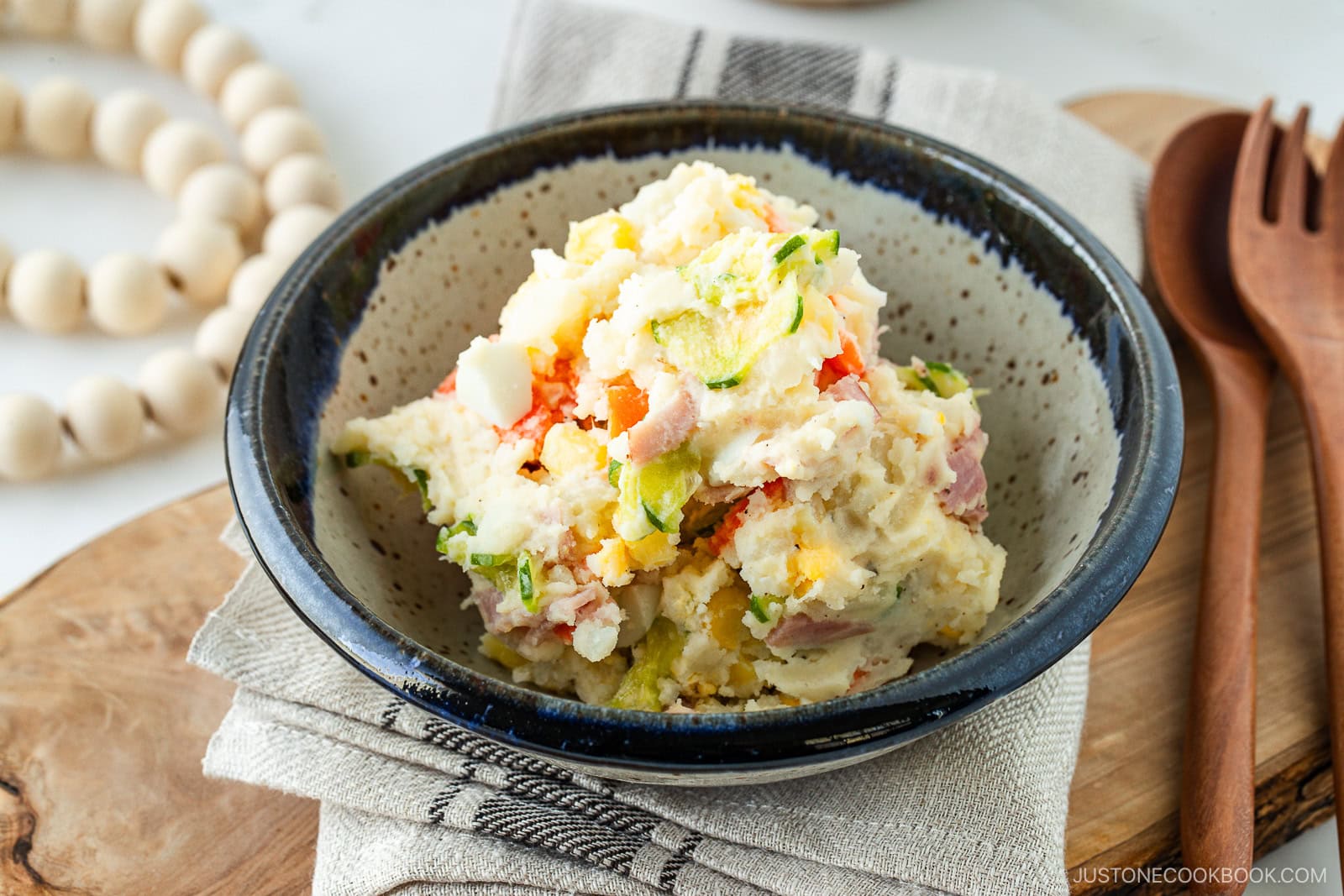 A bowl of Japanese potato salad with visible chunks of potato, carrot, cucumber, and ham, served in a rustic ceramic bowl on a folded cloth napkin beside a wooden fork and spoon.