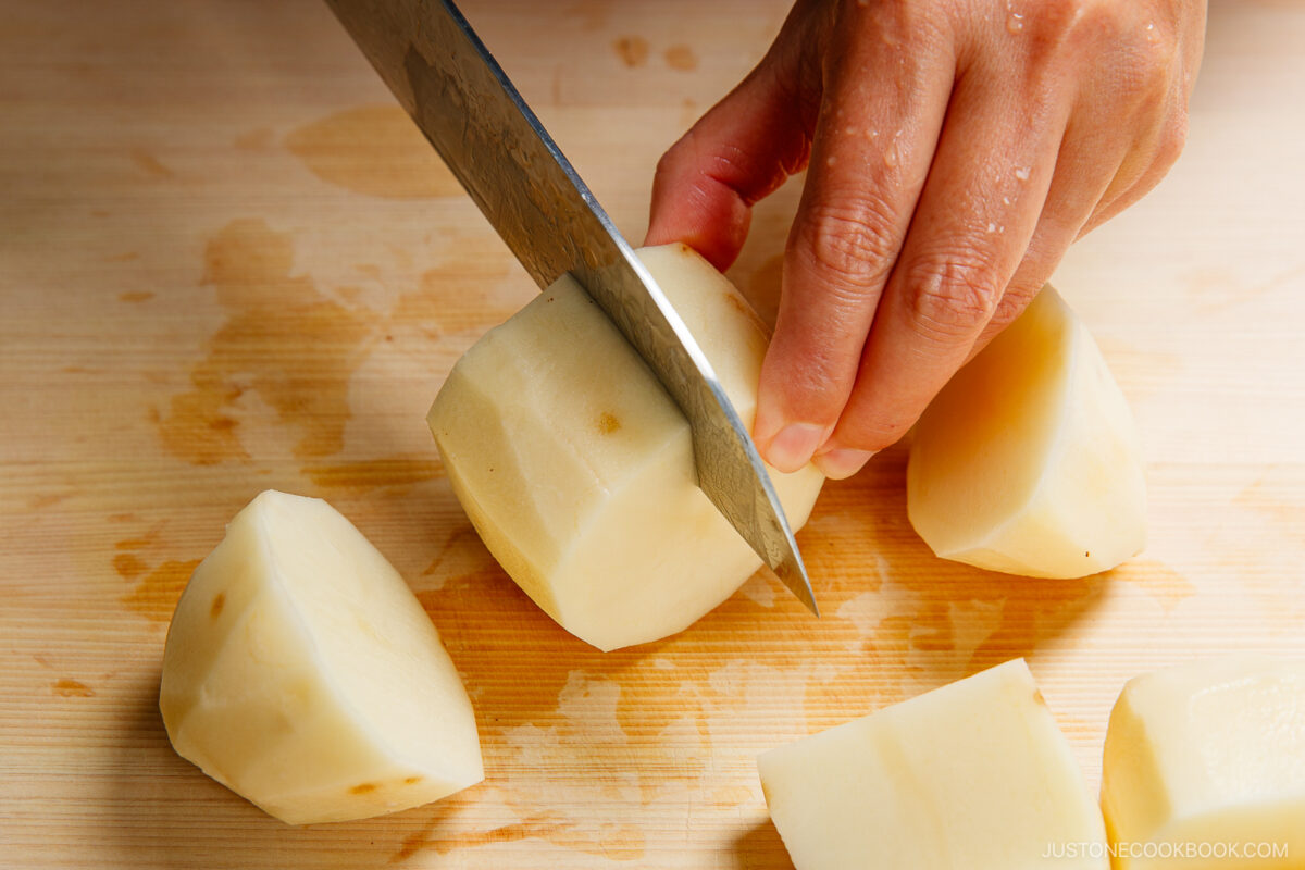 A hand slices a peeled potato with a large knife on a wooden cutting board, surrounded by several chunks of potato.