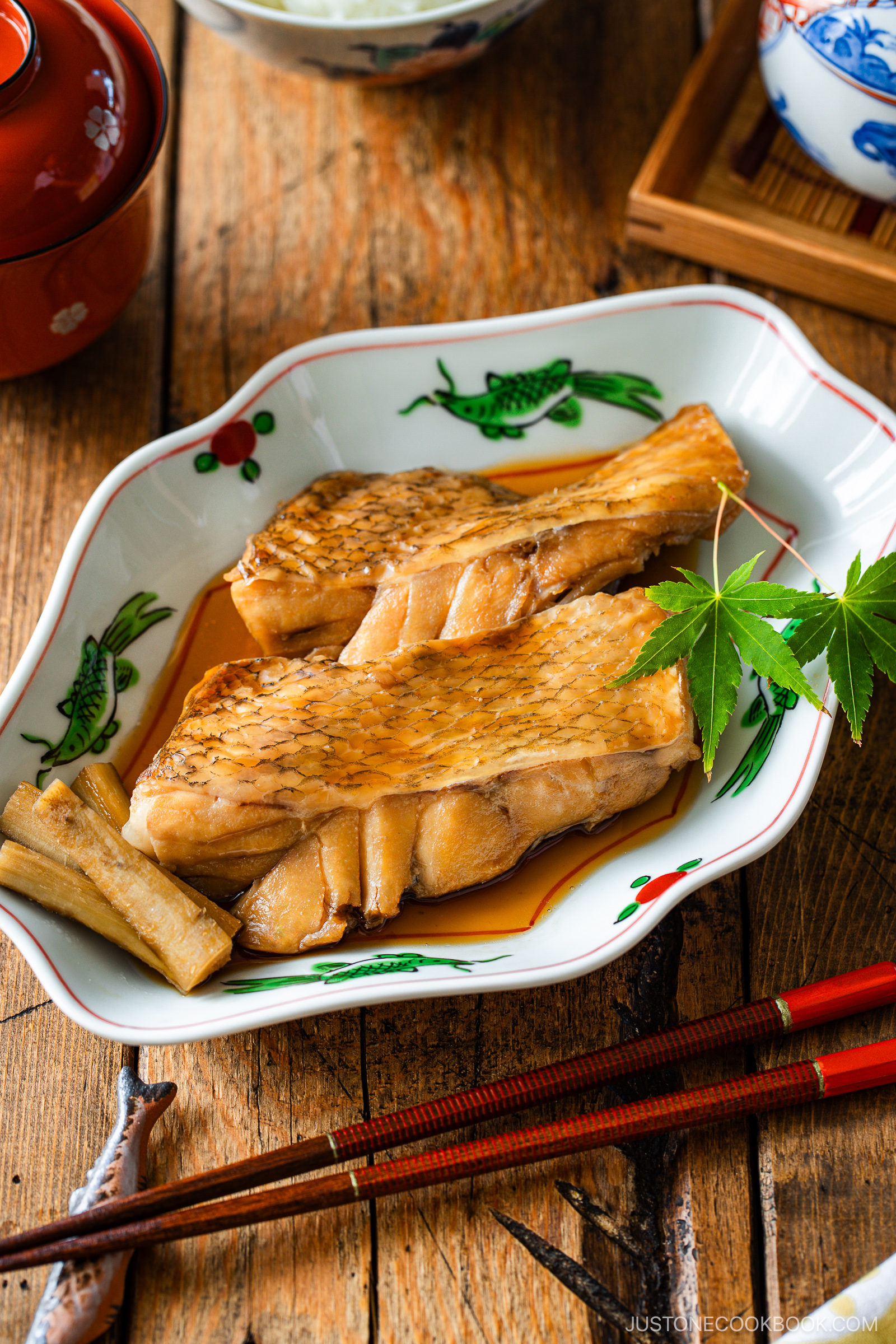 Two pieces of simmered fish served in a decorative dish with a green maple leaf garnish, placed on a wooden table alongside chopsticks and other small dishes.
