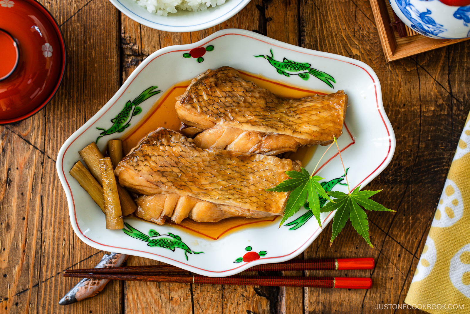 A plate of simmered fish fillets in soy-based sauce, garnished with a green maple leaf and vegetables, sits on a wooden table with chopsticks, a bowl of rice, and a red miso soup bowl nearby.