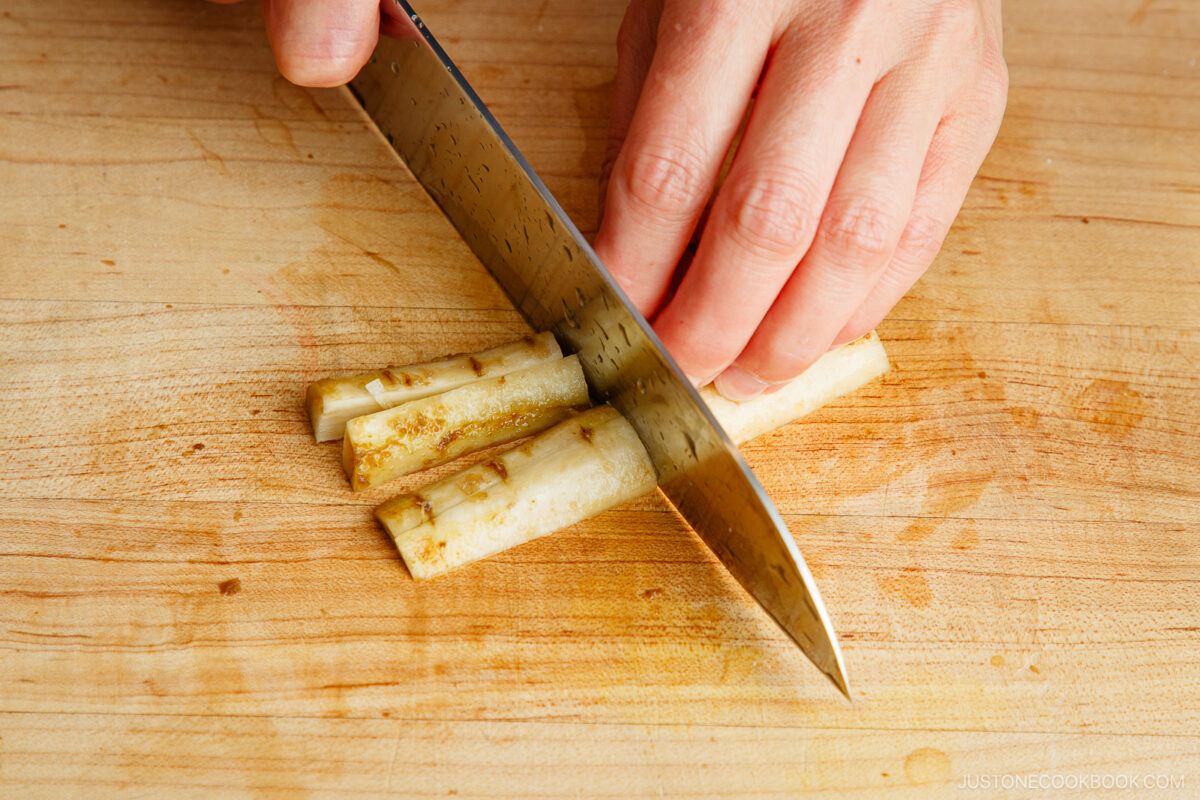 A hand holds a knife and slices burdock root into thin strips on a wooden cutting board.