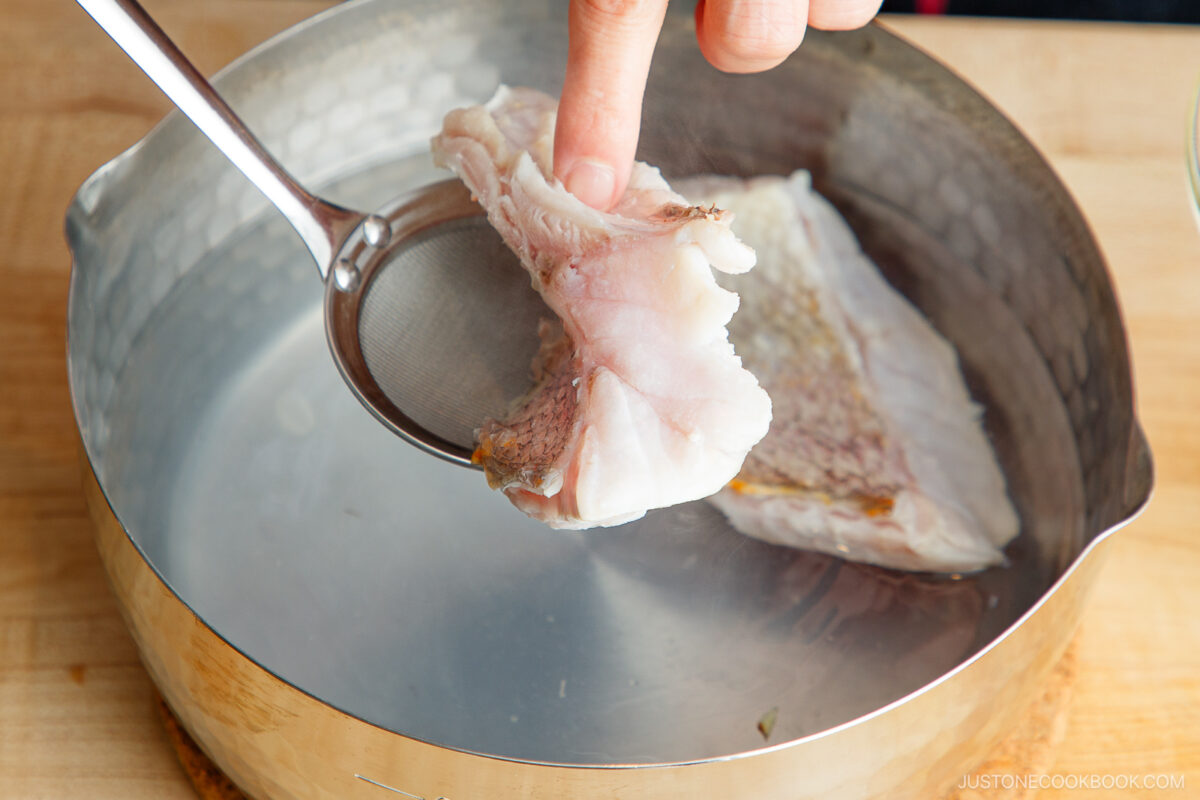 A hand uses a mesh skimmer to hold a piece of cooked fish over a pot of hot water on a wooden surface.