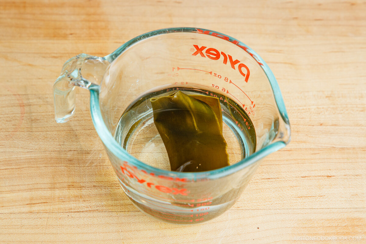 A glass measuring cup with water and a piece of dried kombu seaweed soaking inside, placed on a wooden surface.