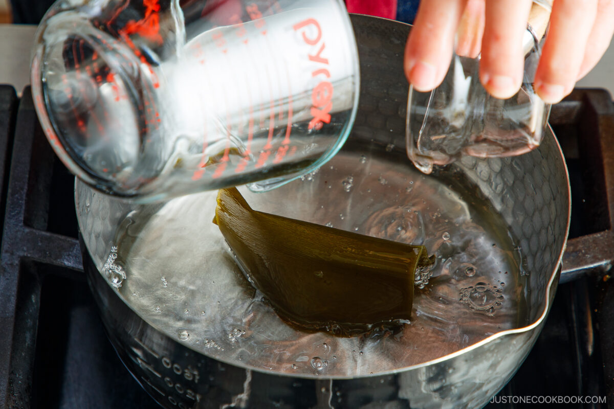 A hand pours liquid from a measuring cup into a pot of water containing a piece of kombu (seaweed) on a stove. Another small glass container is held nearby.