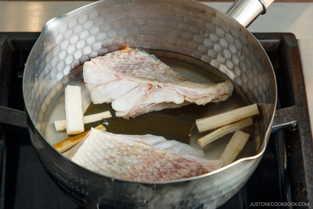 Two raw fish fillets and several burdock root sticks simmer in a metal pot filled with water on a stovetop.