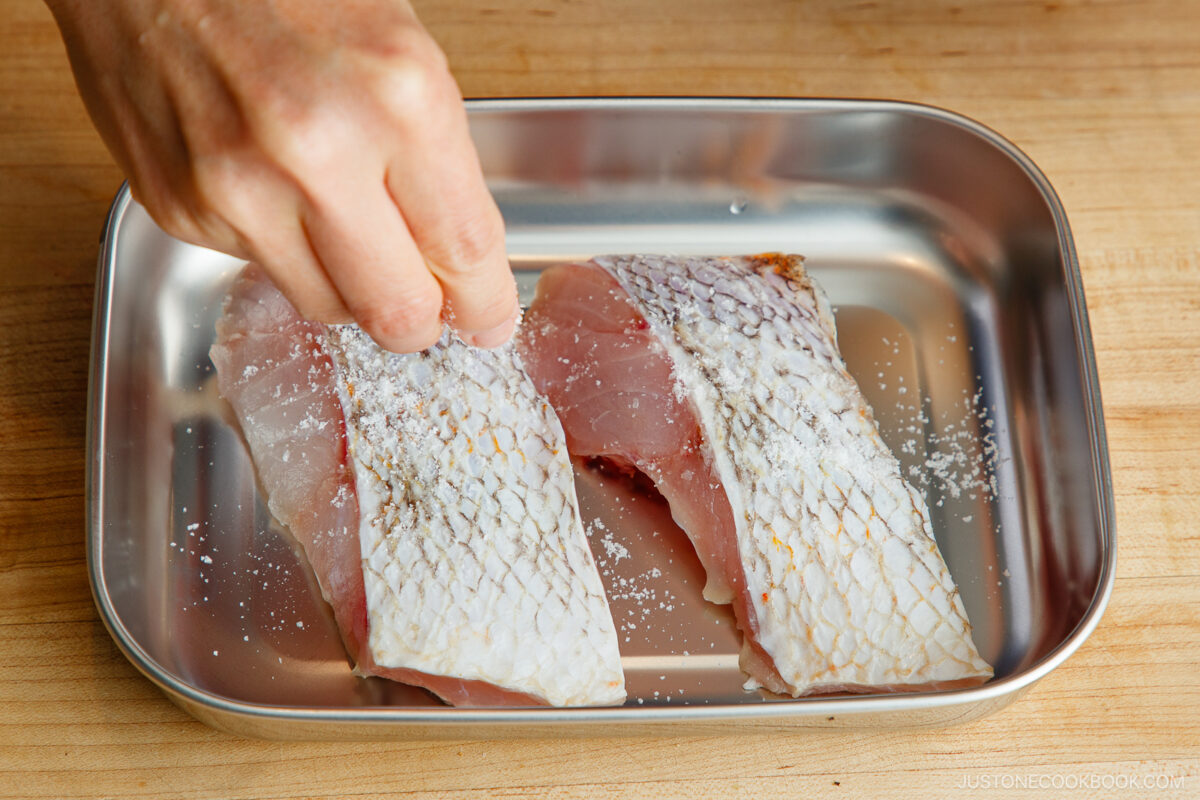 A hand sprinkles salt over two raw fish fillets with skin on, placed in a metal tray on a wooden surface.