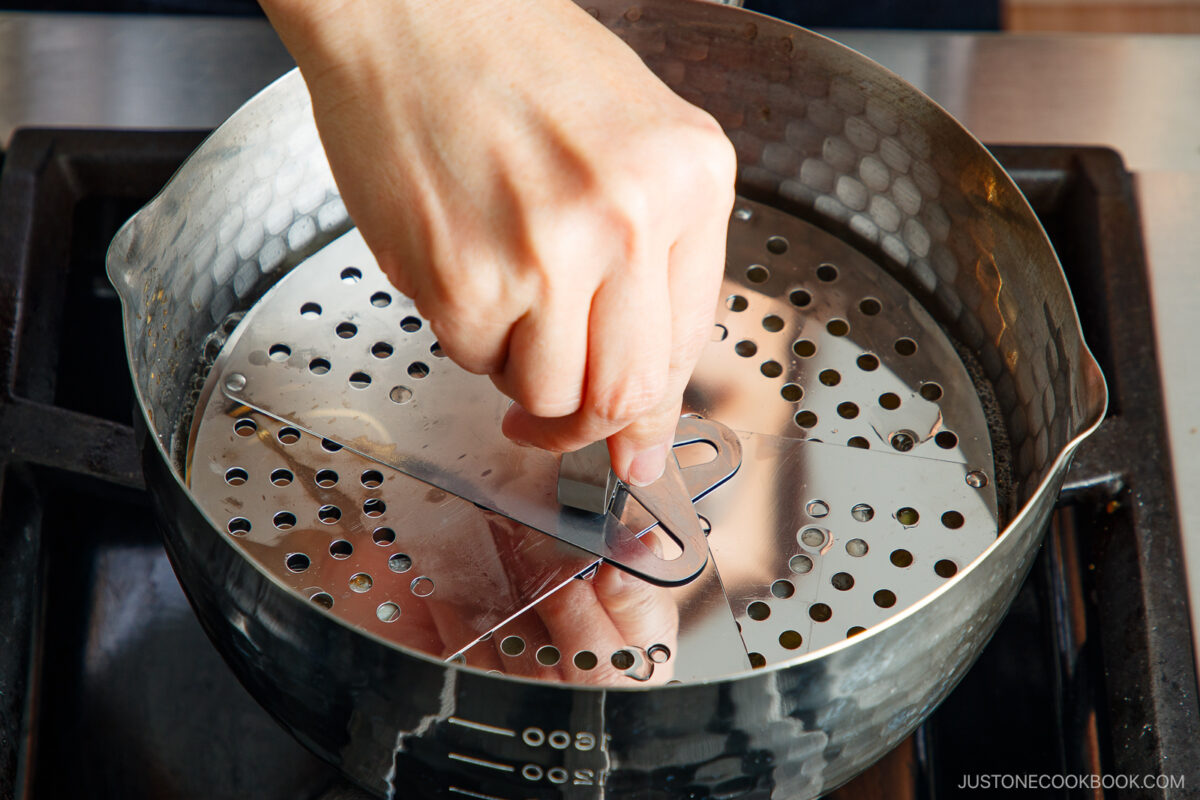 A hand places a metal steamer lid with holes onto a pot on a stovetop, preparing to steam food.