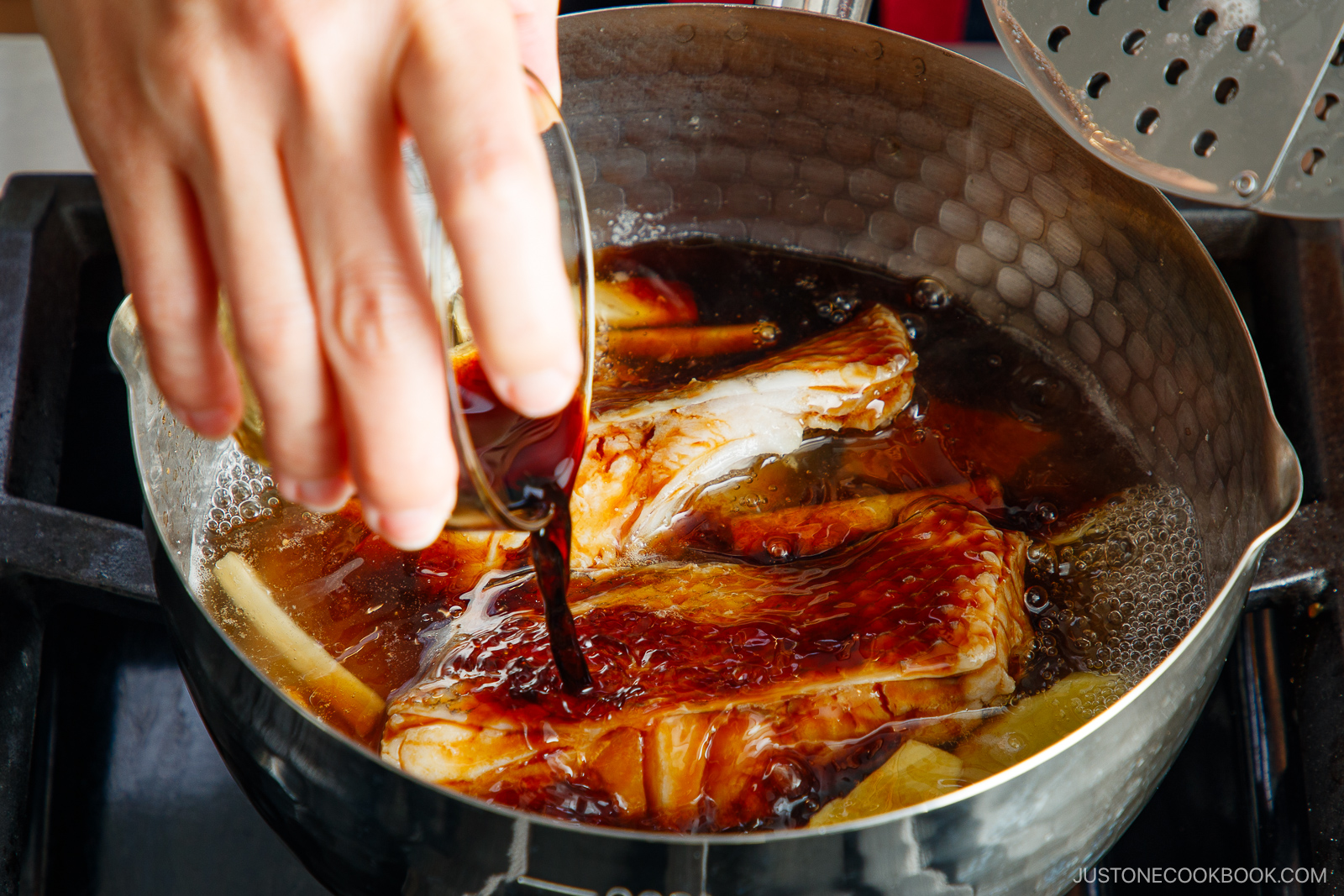 A hand pours dark sauce into a pot containing simmering fish pieces and slices of ginger, preparing a dish on a stovetop.