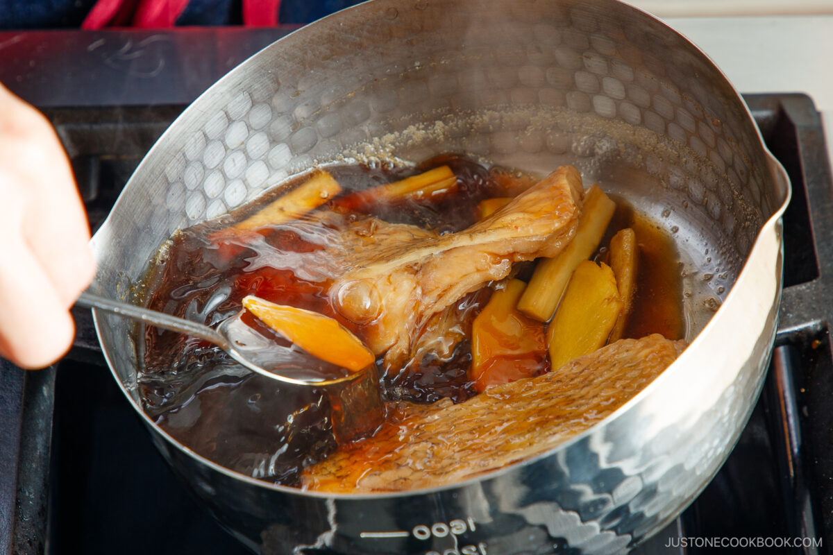 A close-up of a pot on a stove with simmering fish fillets, ginger slices, and green onions in a dark broth. A hand holds a ladle, scooping the broth over the fish. Steam rises from the cooking pot.